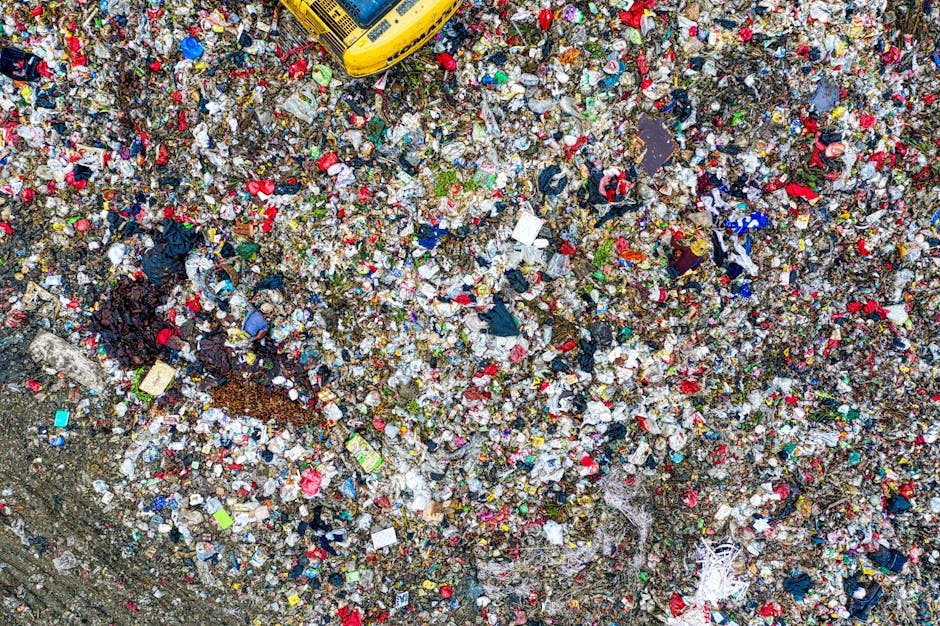 An aerial view of a large landfill site filled with a dense layer of mixed waste materials, including plastic bags, cardboard, food packaging, textiles, and miscellaneous debris, all scattered across 